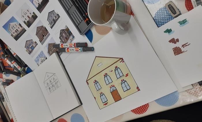 Table with drawing activity happening on it. Table has spotty tablecloth upon which there is a poster of chapel photographs by Paul Cabuts, an open sm
